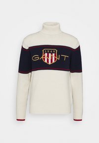 Crème kleurige trui met een hoge hals, voorzien van marineblauwe horizontale strepen en een schildlogo met rode en gouden accenten. Zachte textuur, casual pasvorm.