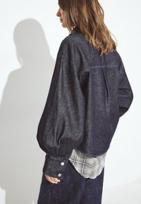 Tmavomodrá džínová košeľa s naberanými rukávmi, gombíkovými manžetami a kockovaným tričkom viditeľným na okrajoch. Hladká textúra s voľným strihom.