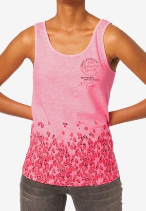 Kobieta ubrana w różowy top bez rękawów z kwiatowym wzorem na dole i napisem na górnej lewej stronie klatki piersiowej, w połączeniu z ciemnymi dżinsami.
