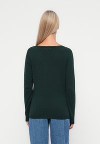 Grön texturerad tröja med långa ärmar och vid halsringning, tillsammans med blå denimjeans. Detaljerat bikake-mönster på tyget.