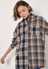 Plaid button-up shirt featuring a mix of beige, navy, and gray colors with a chest pocket and frayed hem. Made from flannel fabric.
