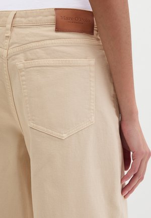 Beige High-Waist-Jeans mit einer Gesäßtasche und einem braunen Lederetikett der Marke Marc O'Polo, getragen von einer Person mit locker am Körper hängender Hand.