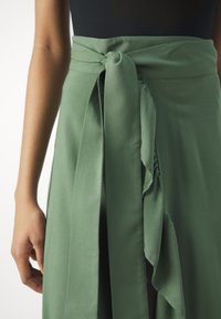 Gonna di tessuto verde con cintura annodata e design fluido. La texture appare liscia con una finitura simile alla seta e dettagli di drappeggio sottili.