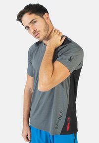 Camiseta gris de manga corta con acentos negros, cuenta con un bolsillo con cremallera en el costado y un logotipo en la parte superior del brazo. Textura suave.