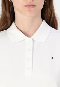 Bílá polo košile s texturovaným povrchem, klasickým límcem a pěti knoflíkovou légou. Na hrudi je výrazné malé logo v červené, bílé a modré barvě.