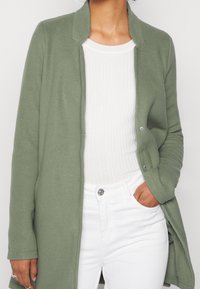 Donna che indossa un cappotto aperto verde chiaro sopra una maglietta bianca a coste e pantaloni bianchi, con una mano in tasca, su uno sfondo semplice.