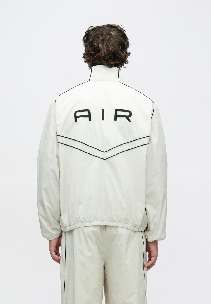 Chaqueta deportiva de nailon blanca con cuello alto, costuras negras de acento y el logo "AIR" en la espalda. Se lleva con pantalones deportivos a juego.