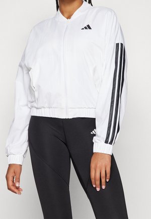 Frau trägt weiße Jacke mit Reißverschluss und schwarzen Streifen an den Ärmeln sowie schwarze Leggings mit Logo am Oberschenkel, steht vor schlichtem Hintergrund.