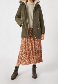 Groene parka met capuchon en een voering van bont, gelaagd over een lange bruine maxi rok met bloemenprint. Donkere laarzen maken de outfit compleet.