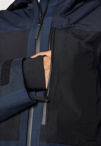 Marineblaue Jacke mit matter Oberfläche, ausgestattet mit einer Reißverschlusstasche auf der Brust, einem geformten Reißverschlussring und strukturierten schwarzen Akzenten.