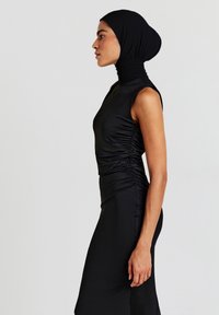 Donna che indossa un abito aderente nero senza maniche con lati arricciati e un foulard nero, in piedi di profilo contro uno sfondo neutro.