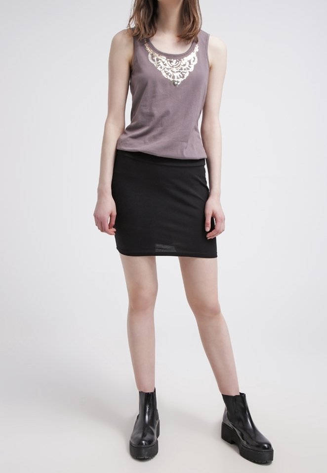 Femme debout portant un haut violet sans manches avec un détail en dentelle blanche, une mini-jupe noire et des bottines noires sur un fond uni.