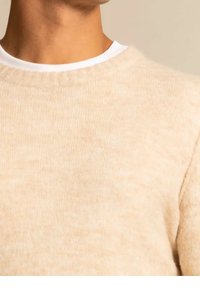 Nahaufnahme von Hals und Schulter, die einen hellbeigen Strickpullover über einem weißen Rundhalshemd tragen, vor einem neutralen Hintergrund.