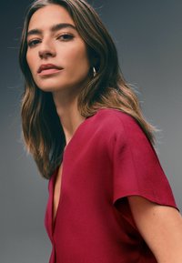 Maroon blouse with a deep V-neck, short sleeves, and a smooth texture, set against a gray background. Subtle stitching detail visible.