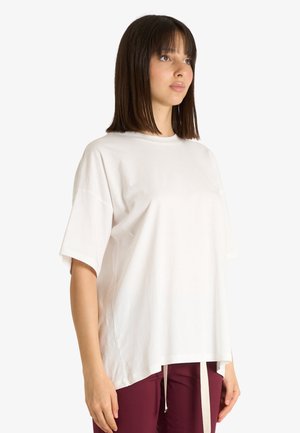 Giovane donna con capelli scuri dritti indossa una camicia bianca larga a maniche corte e pantaloni color bordeaux, in piedi davanti a uno sfondo chiaro e semplice.