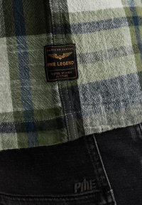 Chemise en carreaux verts et gris avec une texture en lin, arborant une étiquette PME Legend, associée à un jean en denim noir avec des broderies de marque.