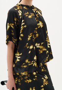Blouse à motif floral noir et doré avec un col froncé et des manches courtes, fabriquée en tissu léger, présentant une silhouette ample.