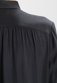 Czarna koszula z kołnierzykiem, o gładkiej fakturze, z marszczeniami z tyłu, które dodają kształtu i elastyczności.