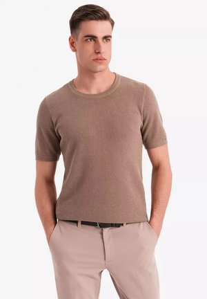Suéter de punto beige de manga corta con cuello redondo y patrón de tejido texturizado, combinado con pantalones de color claro y un cinturón negro.