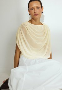 Top drappeggiato di colore crema realizzato in tessuto trasparente abbinato a una gonna bianca. Accenti notevoli includono orecchini importanti. Modella seduta su uno sfondo bianco.