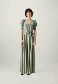 Hosszú, nőies, könnyed ruha puha zöld anyagból, rövid, fodros ujjakkal és övvel a deréknál. Mély V-kivágása van.