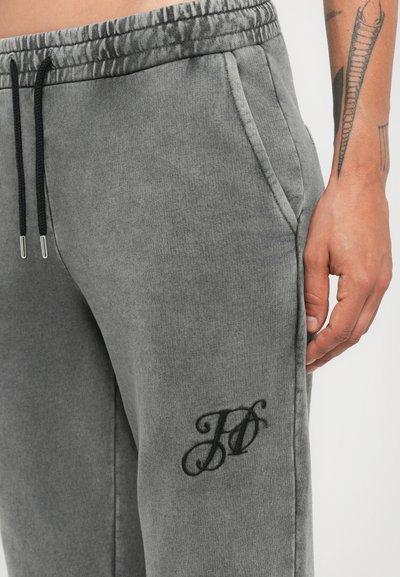 Close-up van een persoon in grijze joggingbroeken met zwarte trekkoorden en een geborduurd "JD" logo nabij de voorkant van de zak, waar een getatoeëerde onderarm zichtbaar is.