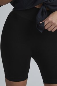 Shorts noirs en stretch avec une large ceinture, texture lisse et coupe au niveau du genou, portés avec un haut sombre noué.