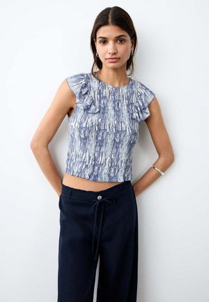 Jonge vrouw draagt een blauw patroon gerimpelde mouwloze top en zwarte broek met trekkoord, staat met handen achter haar rug.