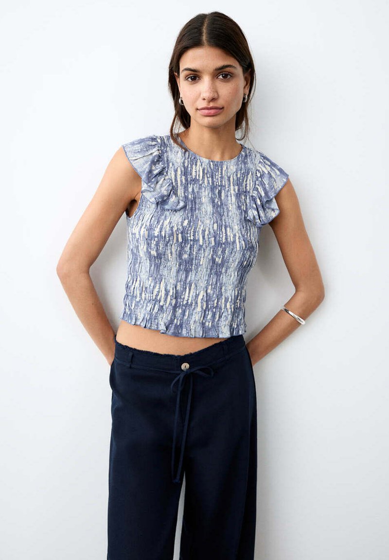 Jeune femme portant un haut sans manches à volants bleu à motifs et un pantalon noir avec cordon, debout les mains dans le dos.