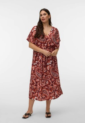 Moteris ilgais tamsiais plaukais, vilkinti raudoną gėlių rašto midi suknelę ir juodus atvirus aukštakulnius, stovinti priešais baltą foną.