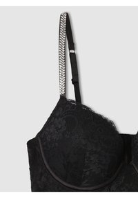 Reggiseno in pizzo nero con coppe sagomate, spalline regolabili e una cinghia decorativa con accenti argentati. Presenta motivi floreali e una texture liscia.