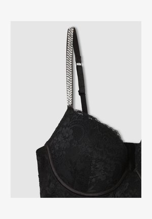 Reggiseno in pizzo nero con coppe sagomate, spalline regolabili e una cinghia decorativa con accenti argentati. Presenta motivi floreali e una texture liscia.