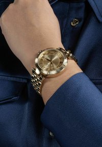 Guldtonad armbandsur med texturerad urtavla, romerska siffror och en länkad metallarmband, visad på en handled mot en blå skjorta.