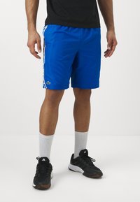 Μπλε αθλητικά σορτς με λευκές ρίγες στο πλάι και λογότυπο Lacoste. Φοριούνται με μαύρες παπούτσια και λευκές αθλητικές κάλτσες. Λείο, ελαφρύ ύφασμα.