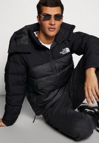 Giacca a vento nera con colletto alto, chiusura con zip e logo, caratterizzata da una finitura opaca e texture trapuntata, abbinata a jeans scuri e occhiali da sole.