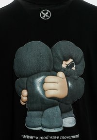 Camiseta negra con un diseño bordado de dos figuras abrazándose, con detalles texturizados y elementos de contraste de color.