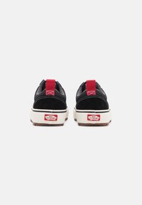 Zapatillas negras de ante con acentos rojos, que cuentan con una entresuela blanca y una suela de goma. La vista trasera muestra la etiqueta del logo en el talón.