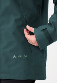 Giacca impermeabile verde scuro con cerniera laterale, polsini regolabili e logo "Vaude" stampato vicino all'orlo. Texture del tessuto liscia.