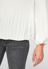 Blusa bianca plissettata con maniche morbide e leggermente a sbuffo; texture liscia; indossata con jeans grigio scuro a vestibilità slim.