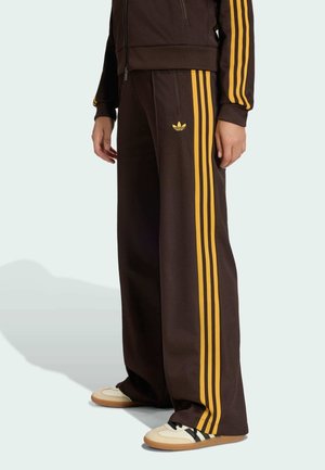 Person trägt dunkelbraune Adidas-Trainingshose und passende Jacke mit gelben Streifen sowie beige Sneakers und steht vor einem schlichten Hintergrund.