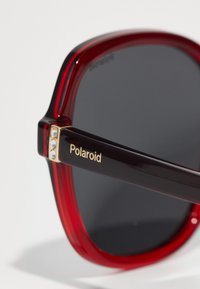 Punase ja mustad prillid läikiva viimistlusega, musta varrega, millel on kuldne "Polaroid" tekst ja kolm väikest sädelust.