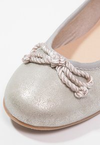 Ballerine en métal argenté avec une surface texturée, dotée d'un bandeau élastique gris et d'un nœud en corde décoratif sur l'orteil.