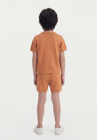 Παιδί με κοντά σκουρόχρωμα μαλλιά, στραμμένο προς τα πίσω, φοράει πορτοκαλί κοντομάνικο μπλουζάκι και ταιριαστά σορτς με λευκά αθλητικά παπούτσια σε απλό φόντο.