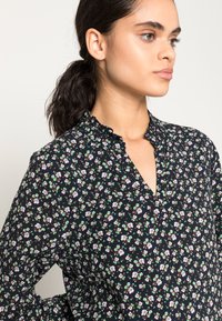 Blusa floral em azul-marinho, com um colarinho franzido e um decote em V. Pequenas flores brancas, verdes e castanhas criam um padrão delicado.