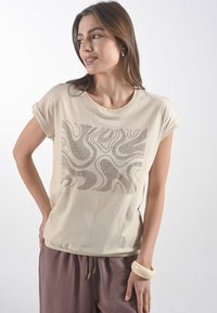 Beige bavlněné tričko s texturovaným grafickým designem v tmavě hnědé barvě, s ohrnutými rukávy a volným střihem. V kombinaci se širokými hnědými kalhotami.
