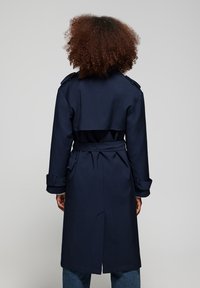 Osoba o kręconych włosach w ciemnoniebieskim płaszczu trencz z paskiem, widziana od tyłu na jednolitym tle.