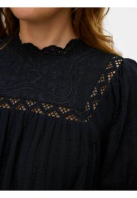 Črna bluzica z izvezenim ovratnikom in čipkastimi detajli. Ima teksturirano tkanino z nabranimi podrobnostmi in valovitim ovratnikom.