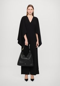 Combinaison noire avec un décolleté en V profond et des manches cape, associée à un grand sac hobo en cuir noir et des escarpins à bout pointu. Texture lisse.