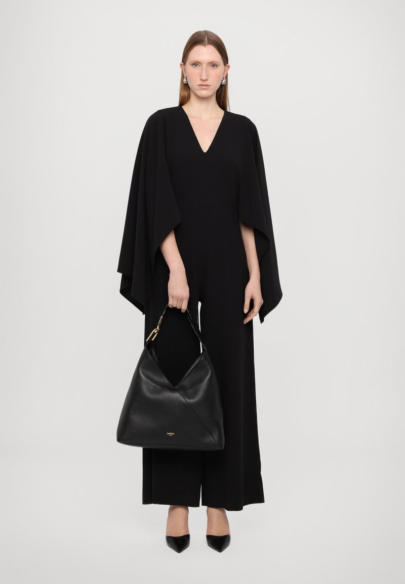 Combinaison noire avec un décolleté en V profond et des manches cape, associée à un grand sac hobo en cuir noir et des escarpins à bout pointu. Texture lisse.