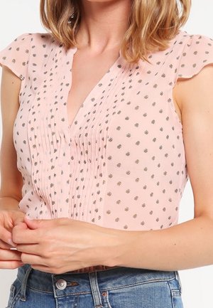 Kvinde iført en lyserød ærmeløs bluse med små blomsterprint og plisseringer, kombineret med blå denimjeans.